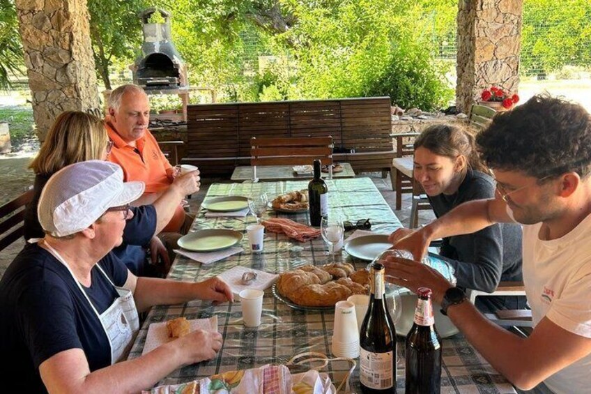 A group of people dining outdoors at a table with food, drinks, and a pizza. 2