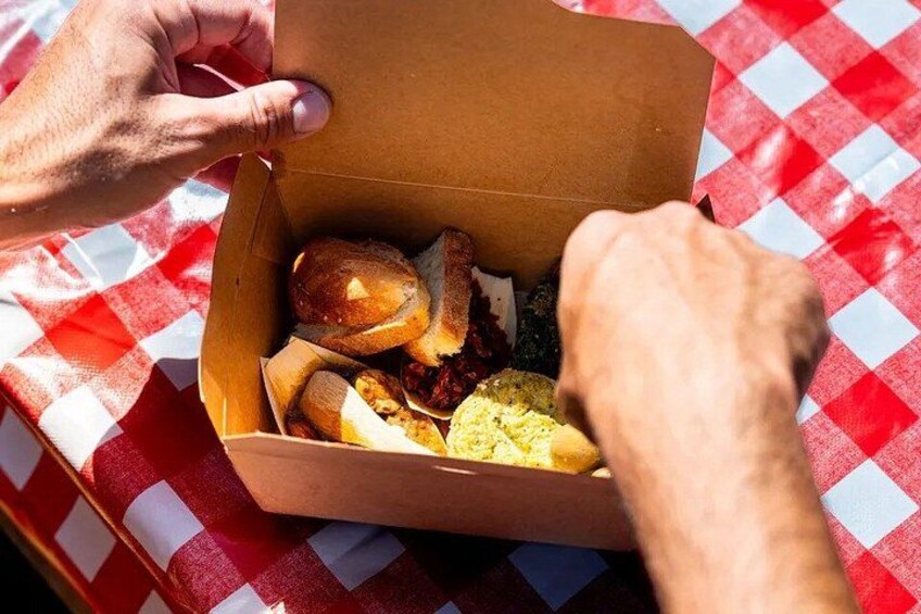 A person opening a cardboard box containing food items on a red and white checkered tablecloth. 4