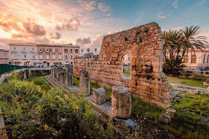 Ancient ruins with stone columns and arches, surrounded by greenery and modern buildings. 1