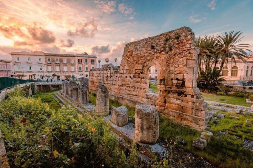 Ancient ruins with stone columns and arches, surrounded by greenery and modern buildings. 1