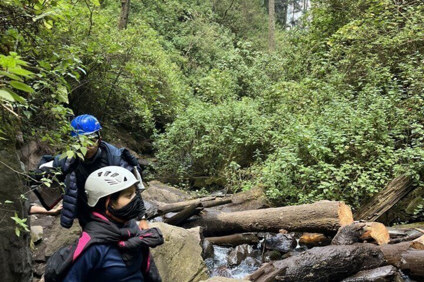 Two people wearing helmets and backpacks near a stream with dense forest in the background. 2