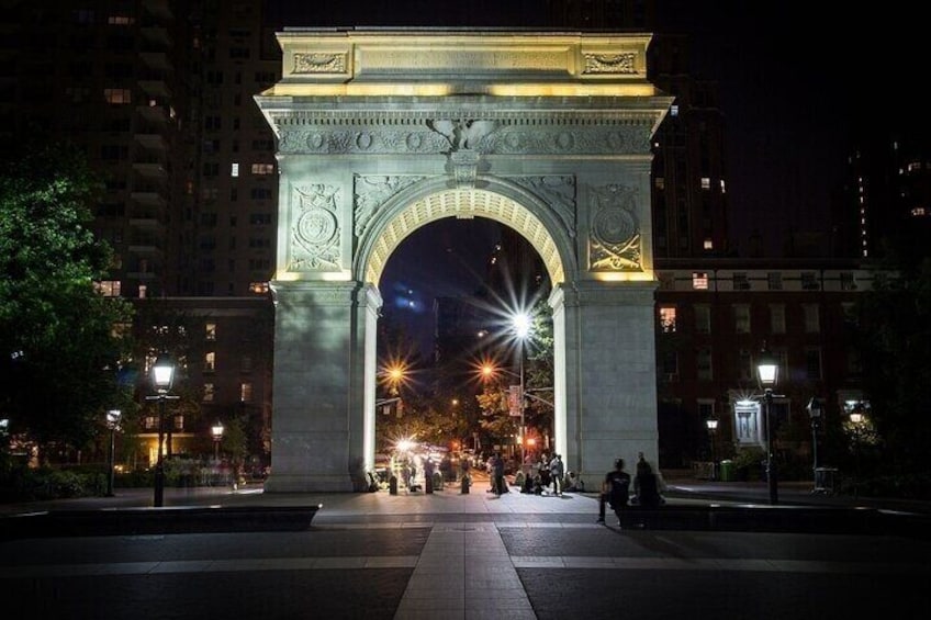 A nighttime view of a large, illuminated archway with intricate carvings, surrounded by a city street with buildings and a few pedestrians. 1
