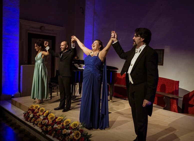 A group of people on a stage with a piano, flowers, and a red chair. 5