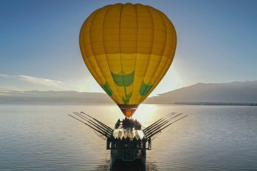 A hot air balloon with a green and yellow design, anchored over calm water with mountains in the background. 1