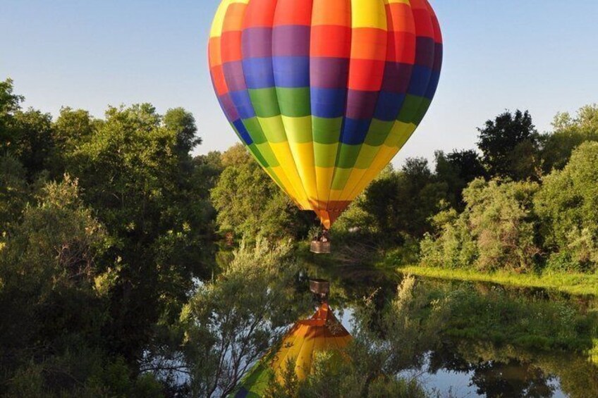 A hot air balloon with a rainbow pattern, flying over a body of water surrounded by trees. 5