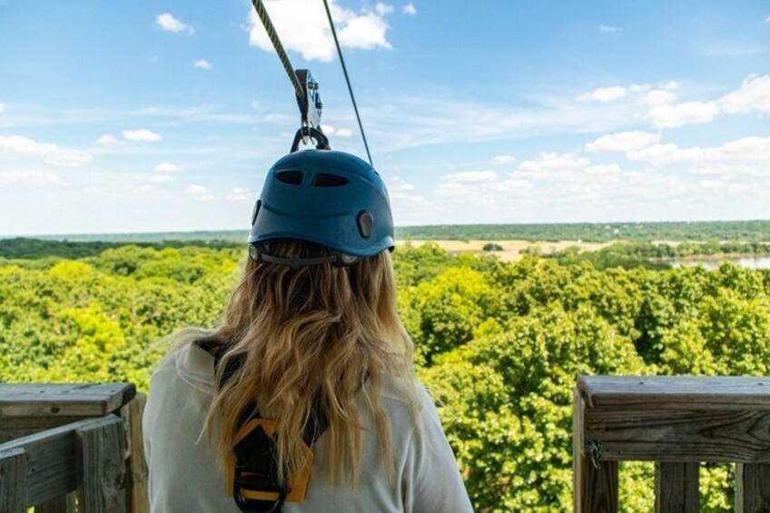 A person with a blue helmet, wearing a white shirt, standing on a wooden platform, overlooking a forested landscape. 4