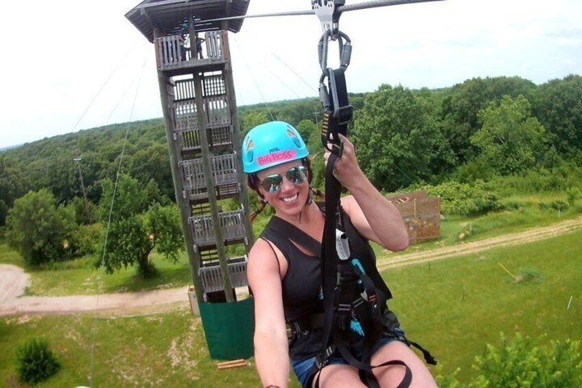 A person in a harness attached to a cable, suspended above a forested area. 1