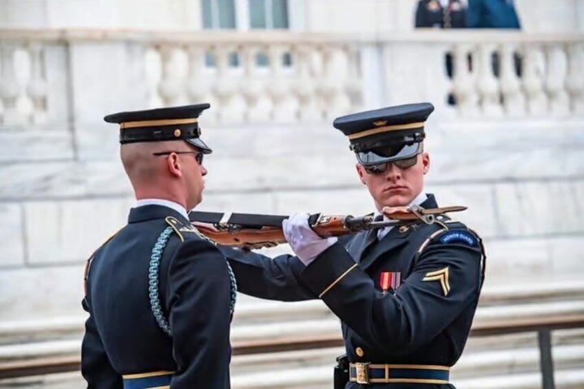 Two uniformed individuals in ceremonial attire, one holding a rifle, standing in front of a marble building with columns. 2
