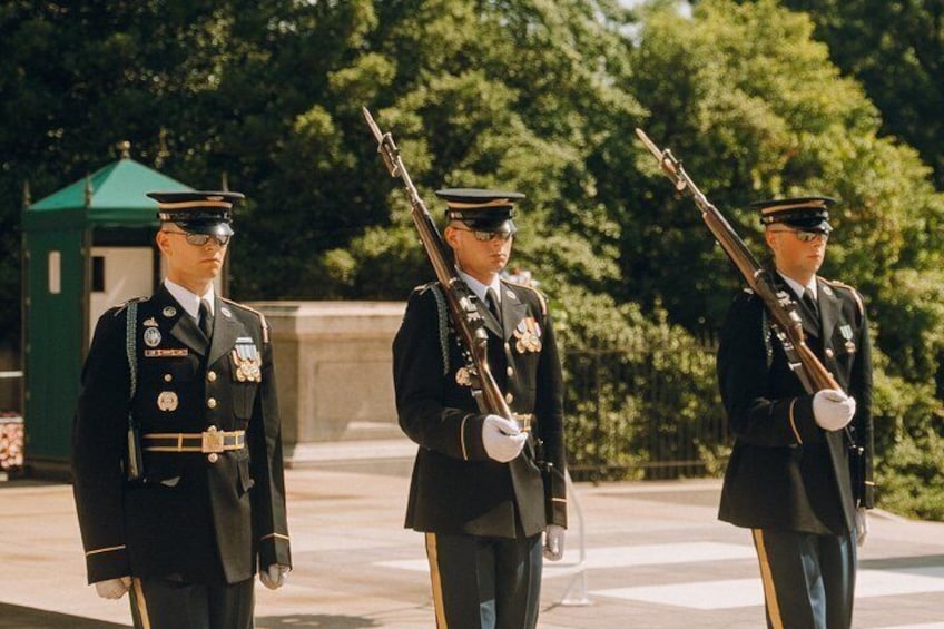 Three uniformed guards standing in a line with rifles, a green structure, and trees in the background. 1