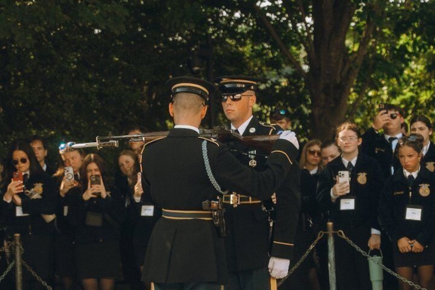 A military ceremony with a uniformed officer in the foreground and a crowd of onlookers behind a rope barrier. 3