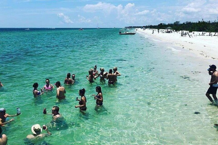 A group of people swimming in clear turquoise water near a sandy beach with boats in the distance. 4