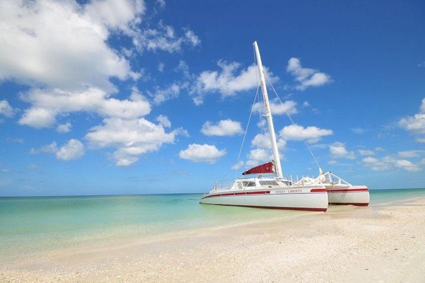 A white and red catamaran named 'Sail Liberty' on a sandy beach with clear turquoise water and a blue sky with scattered clouds. 1