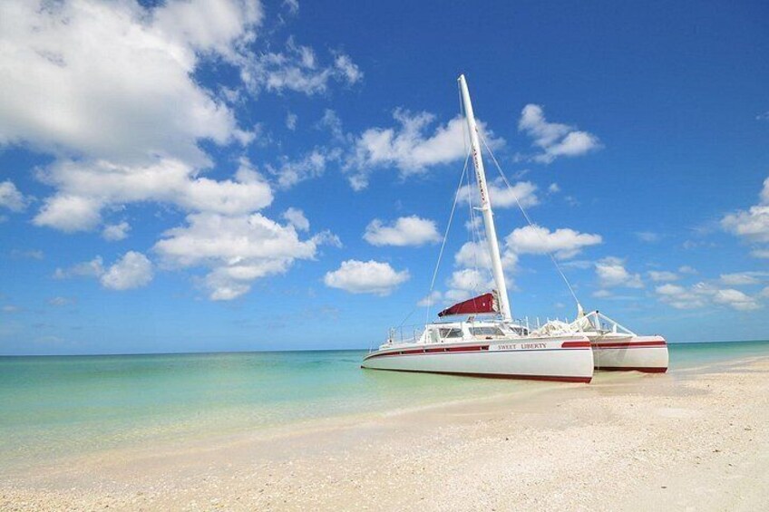 A white and red sailboat named 'Sail Liberty' on a sandy beach with clear turquoise water and a blue sky with clouds. 1