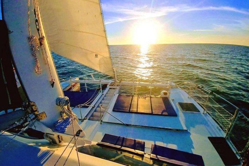 A sailboat with a partially unfurled sail, deck seating, and the sun setting over the ocean. 2