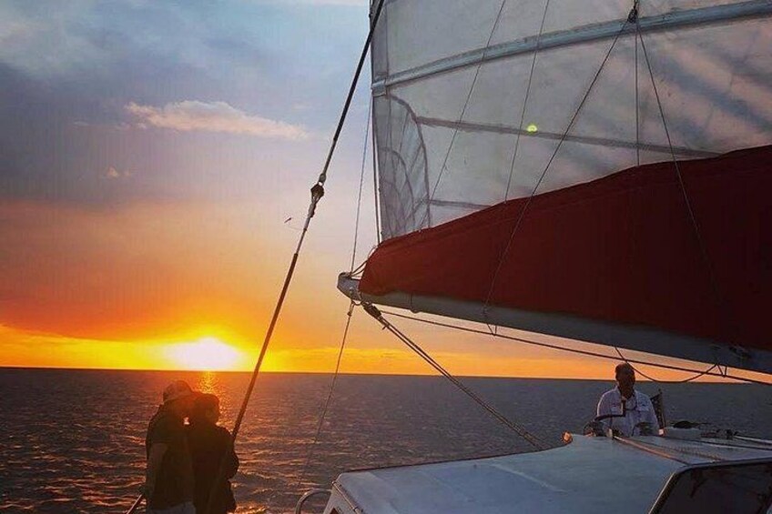 A sailboat with a red sail, a person standing on the deck, and the sun setting over the ocean. 3