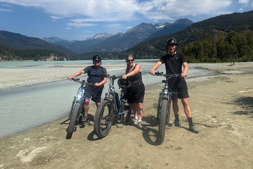 Three people on mountain bikes by a lake with mountains in the background. 1