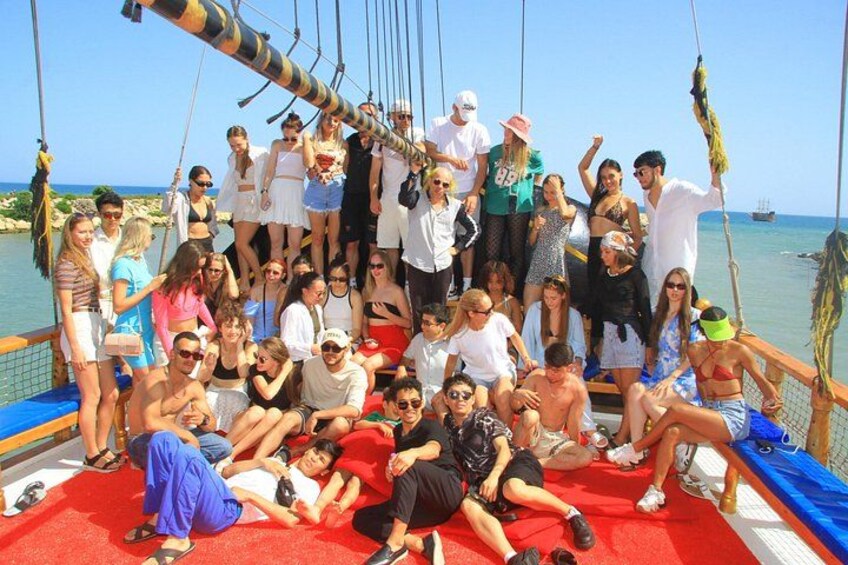 A group of people on a boat with a red deck, some standing and some sitting, with a calm sea and a clear sky in the background. 4