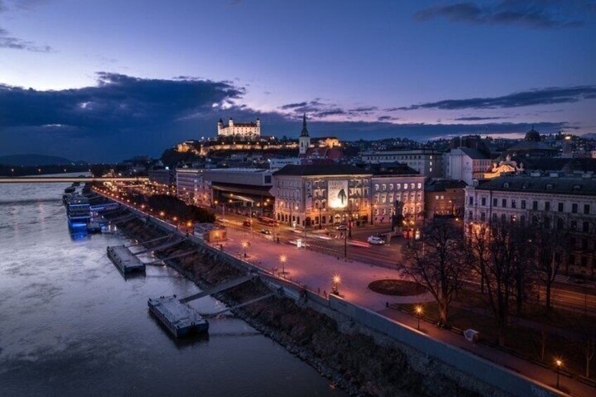 A cityscape at dusk with a prominent castle on a hill, a waterfront promenade, and illuminated buildings. 3