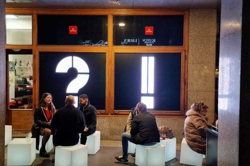 People sitting on white stools in a lobby with large question mark windows. 1