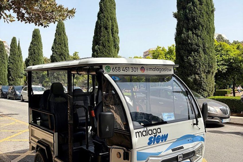 A white and blue bus with 'malaga slow' branding parked in a parking lot with trees and other cars in the background. 2