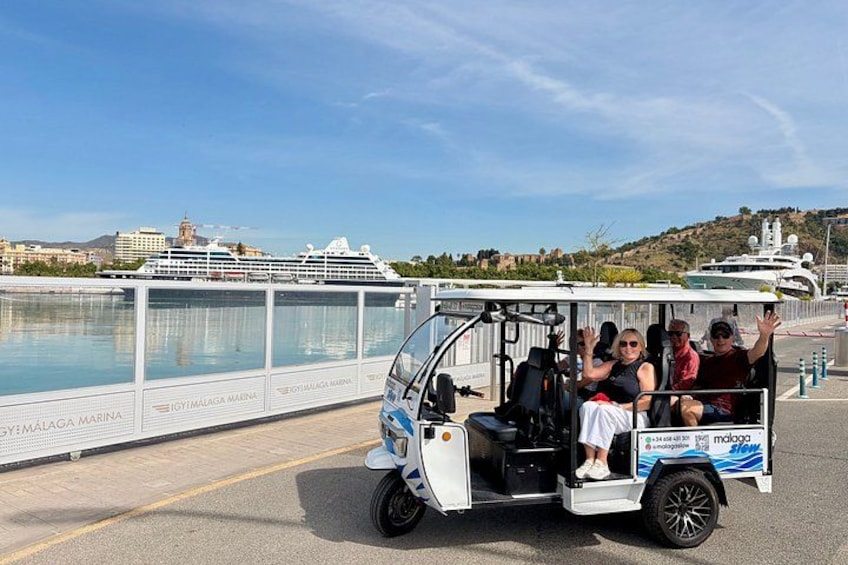 A white and blue golf cart with passengers, a large cruise ship, and a waterfront with buildings in the background. 1
