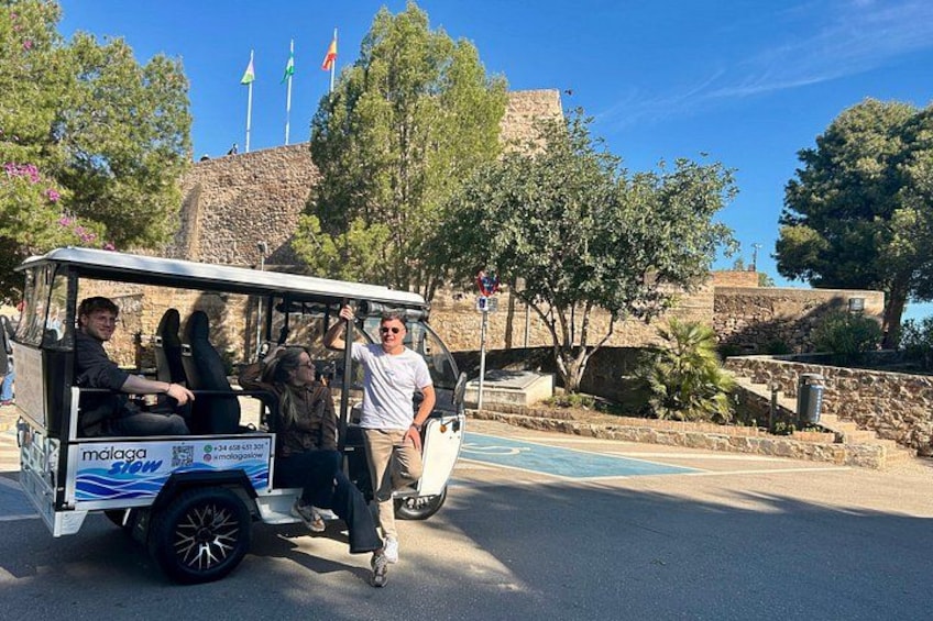 A golf cart with people, a stone wall, trees, and flags in the background. 5