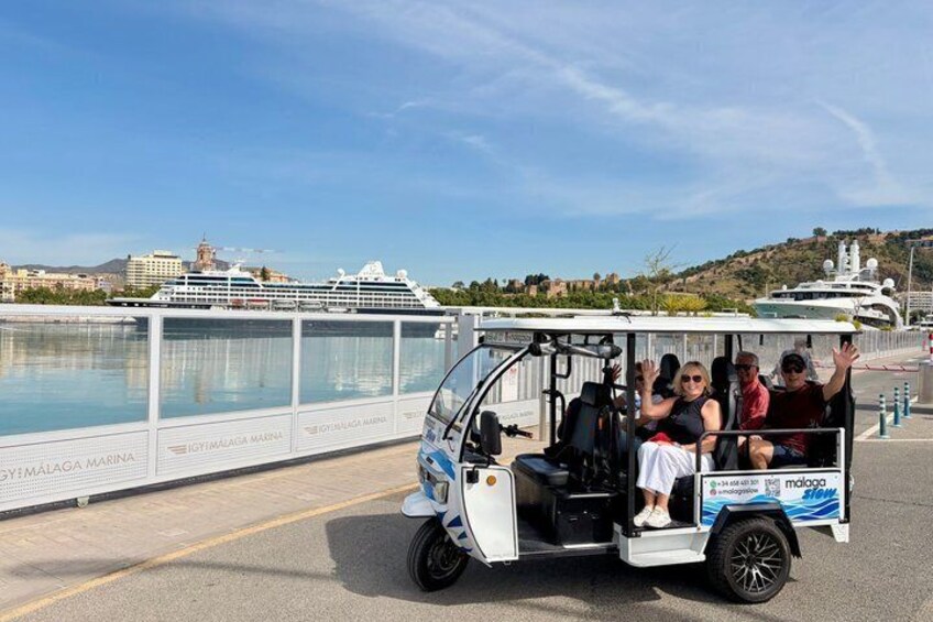 A golf cart with passengers near a waterfront, a cruise ship in the background, and a marina sign. 1