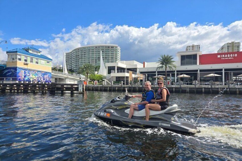Two people jet-skiing on a body of water with buildings and a restaurant in the background. 2