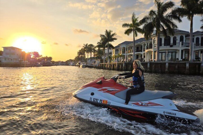 A person jet-skiing on a body of water with a sunset in the background and palm trees along the shore. 1