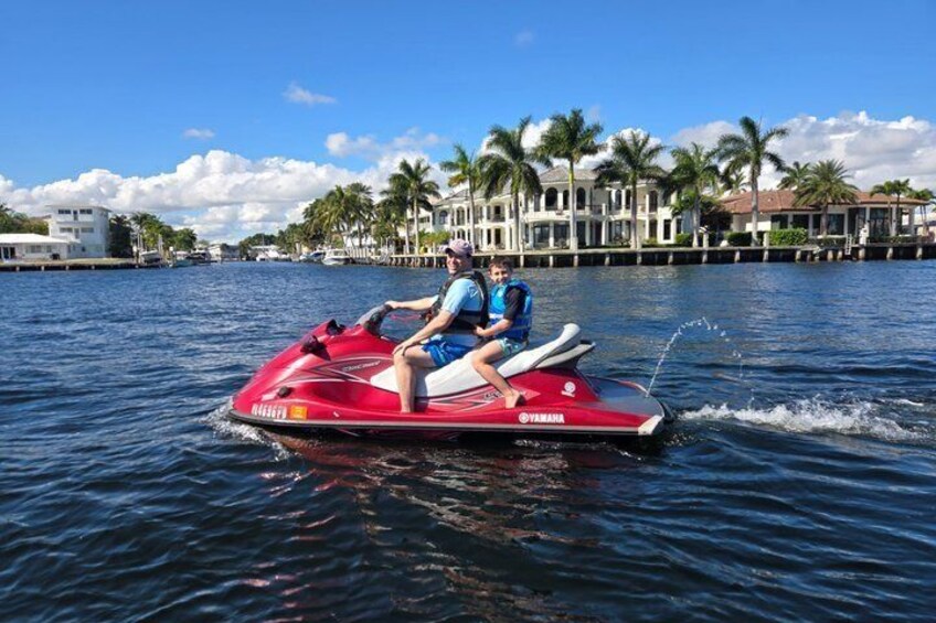 A red Yamaha jet ski on calm water with two people on board, palm trees, and waterfront buildings in the background. 5