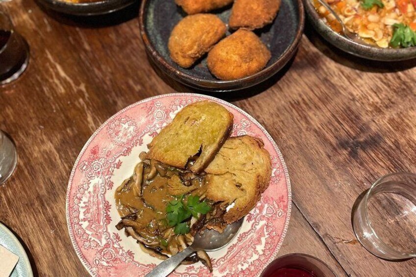 A plate of food with bread and mushrooms, a glass of red wine, and a bowl of soup on a wooden table. 4