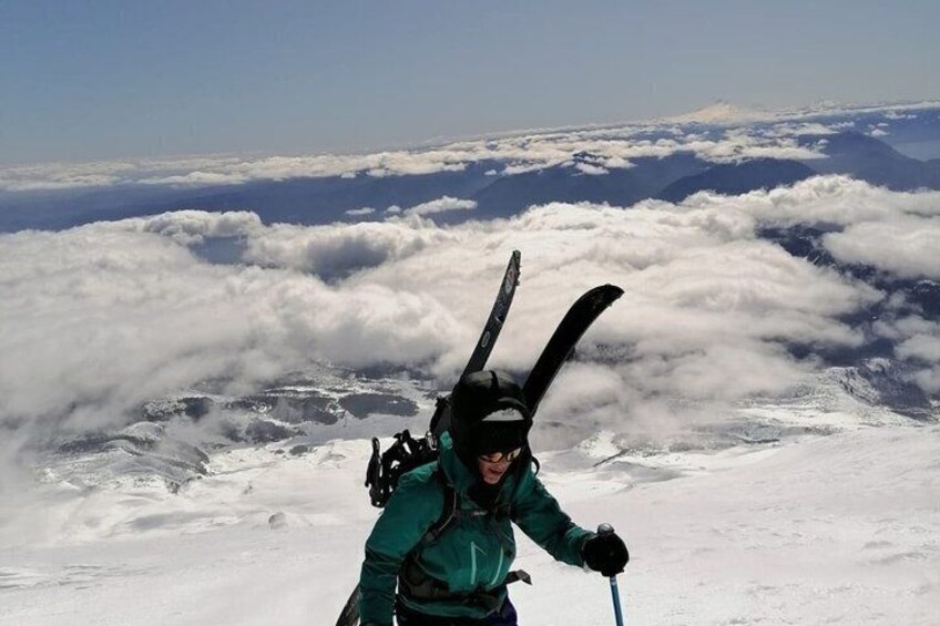 A person in a green jacket with skis strapped to their head, standing on a snowy mountain with clouds below. 4