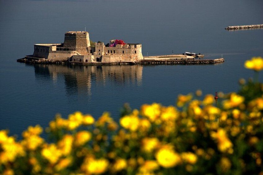 A historic fortification by the water, with a clear sky and yellow flowers in the foreground. 1