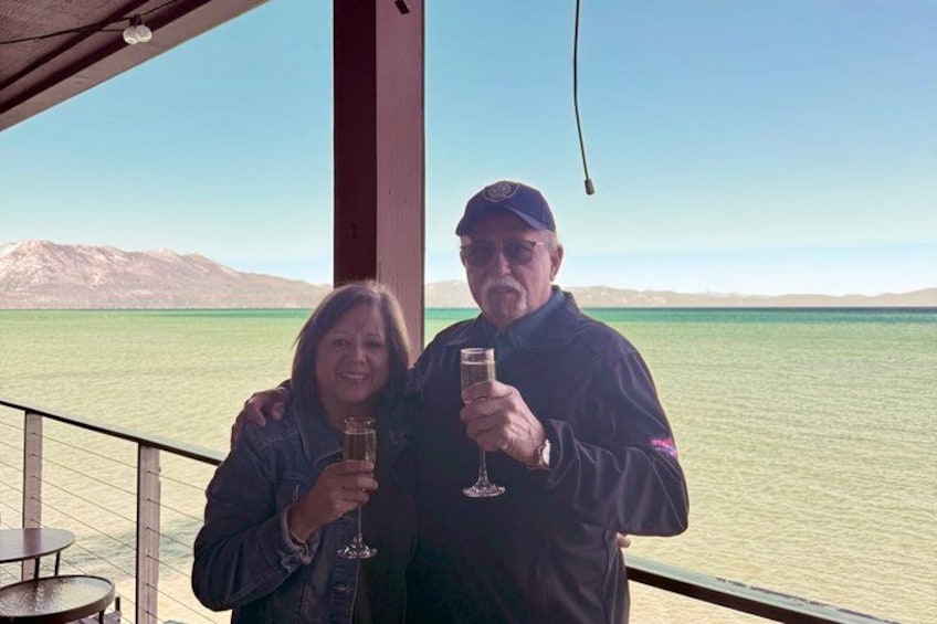 Two people holding glasses on a balcony overlooking a body of water with mountains in the background. 3