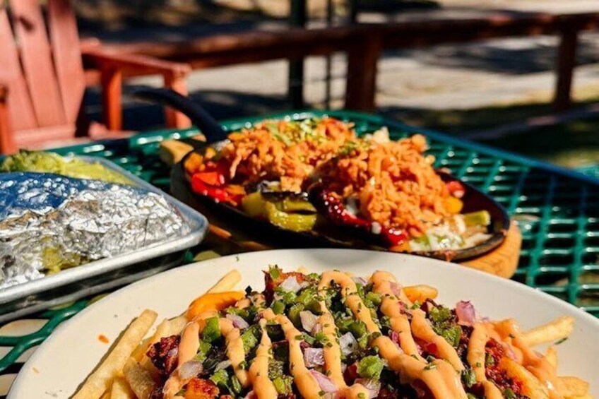 A variety of dishes on a table, including a plate with fries topped with green sauce and onions, a pan with rice and vegetables, and a tray with foil-wrapped food. 5