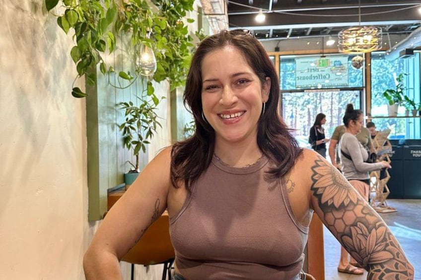 A person seated indoors with a tattoo on their arm, a plant hanging above, and a sign in the background. 5