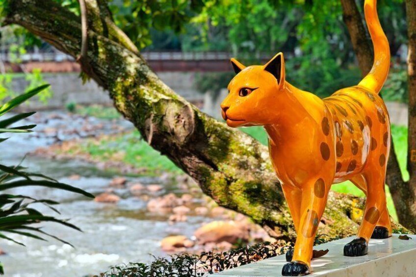 A statue of a cat with spots, standing on a concrete ledge with a river and greenery in the background. 2
