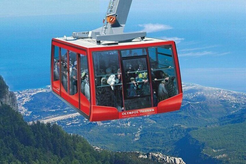 A red cable car with passengers, labeled 'Olympus Teleferik,' suspended above a mountainous landscape with a view of the sea. 1