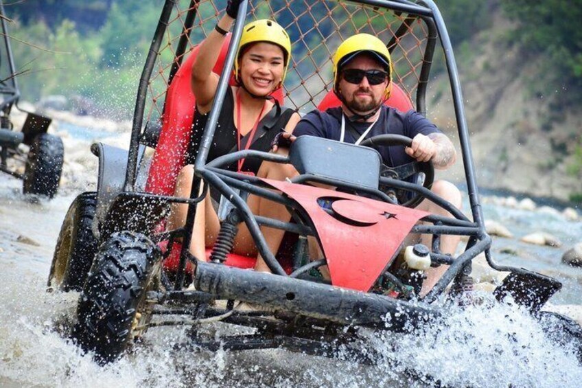 Two people in a four-wheeled vehicle navigating a river with water splashing around. 1