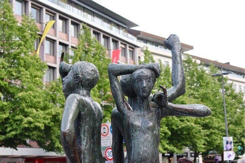 A sculpture of two figures in a public square with buildings and trees in the background. 5
