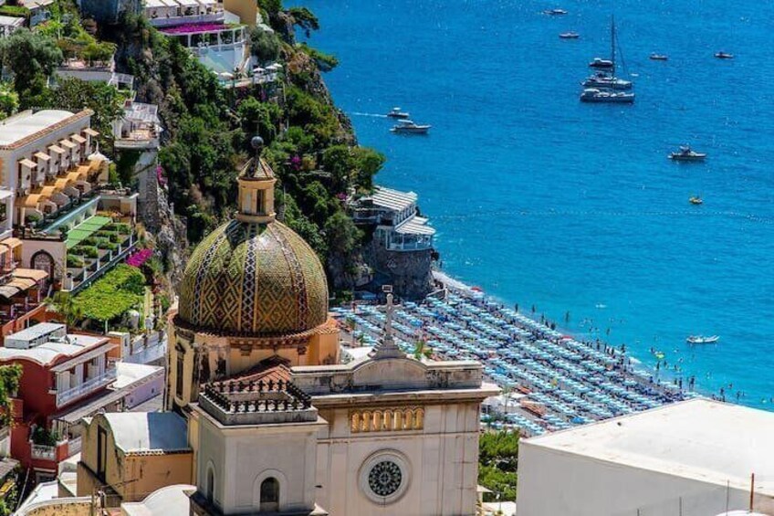 A coastal town with a prominent church dome, a beach with umbrellas, and boats on the water. 1