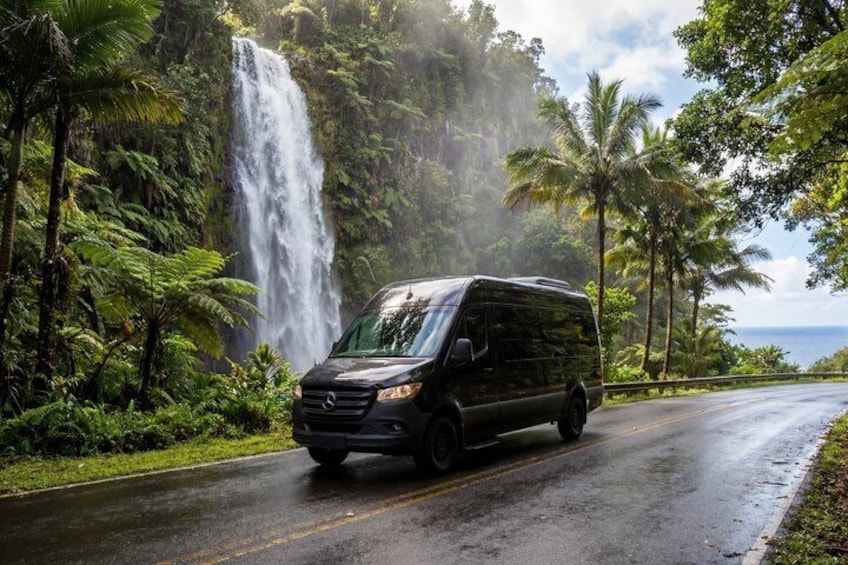 A black van driving on a wet road with a waterfall and palm trees in the background. 1