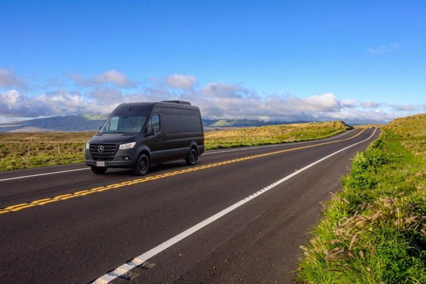 A black van driving on a two-lane road with grassy fields on both sides and a clear blue sky. 2