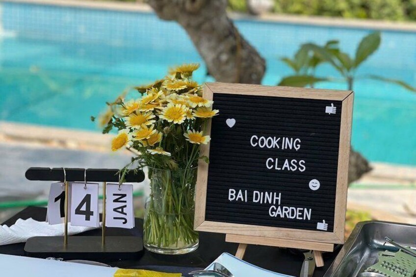 A blackboard sign with 'COOKING CLASS' and 'BAI DINH GARDEN' written on it, a date sign with '14 JAN', and a vase with yellow flowers. 4