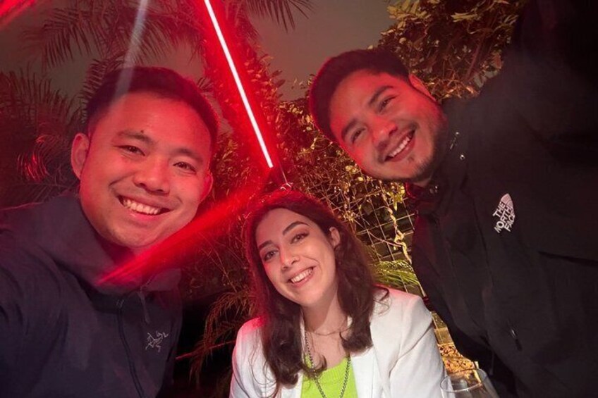 Three people in a dark jacket and a white jacket with a green top, standing in front of a fence with red lighting. 4