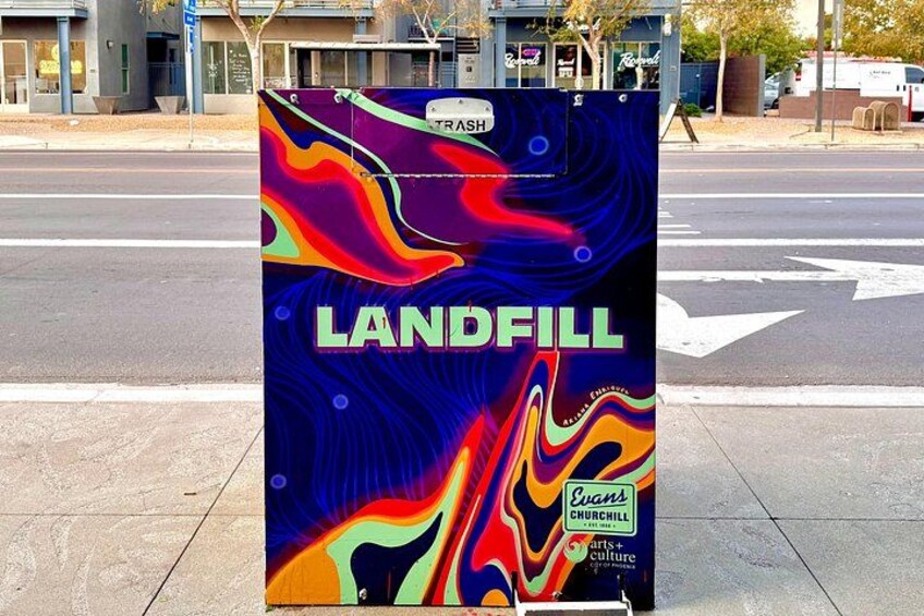 A colorful trash can with the word 'LANDFILL' on it, placed on a sidewalk near a road intersection. 3
