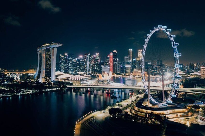A nighttime cityscape with a prominent Ferris wheel, a distinctive modern building with a curved top, and a body of water reflecting the city lights. 2