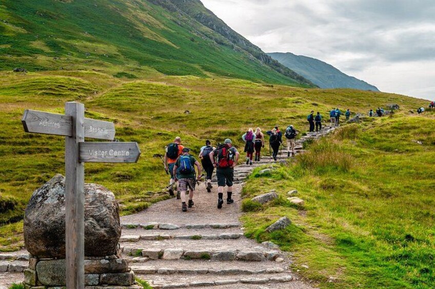 A group of hikers on a stone path in a grassy mountainous area with a signpost indicating directions to 'Visitor Centre' and 'Machinies'. 5