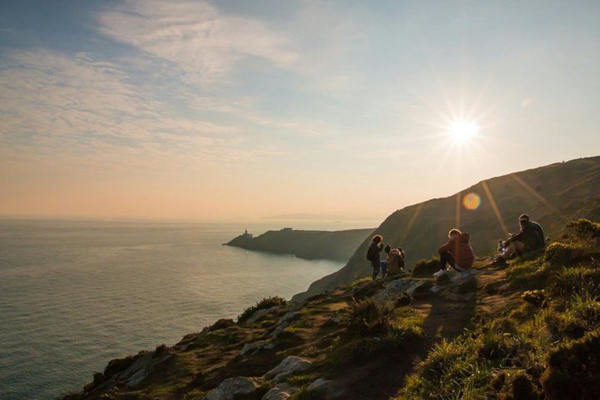A coastal landscape with people sitting on a grassy hill, overlooking the sea and a lighthouse in the distance. 5