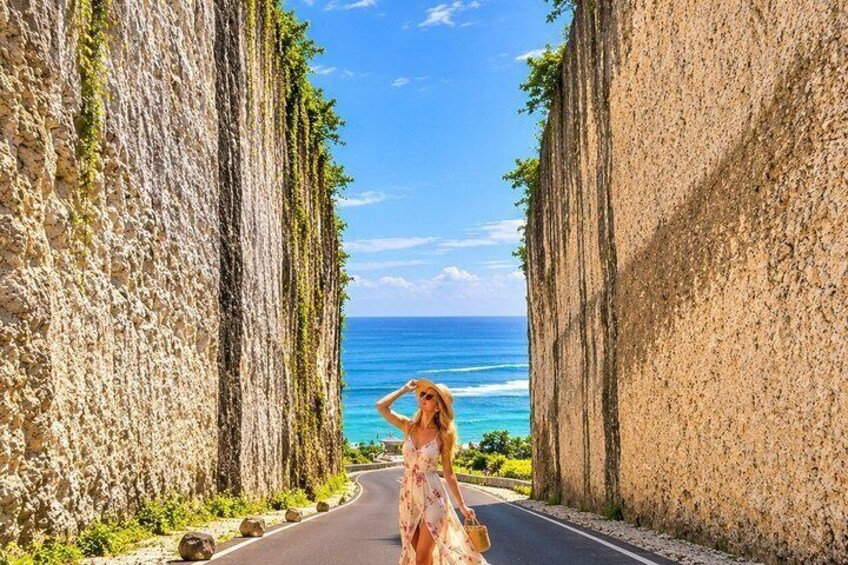 A woman in a dress standing on a road flanked by tall stone walls with a view of the ocean. 2